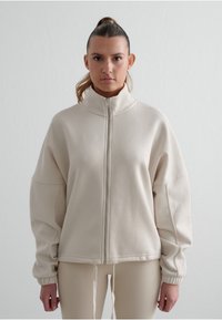 Béžová mikina na zip s vysokým límcem, oversized střihem, spadlými rameny a elastickými manžetami. Hladká textura, minimalistický design.