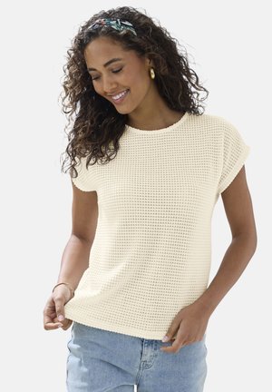 Lächelnde Frau mit lockigem Haar trägt cremefarbenes, kurzärmeliges Oberteil mit Struktur, blaue Jeans, goldene Creolen und ein gemustertes Stirnband.