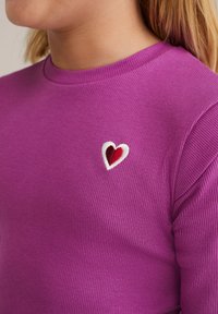 T-shirt à manches longues rayé violet avec un patch en forme de cœur brodé blanc et rouge sur la poitrine. Texture lisse avec un col rond.