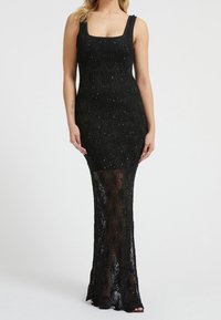 Nainen, joka on pukeutunut hihattomaan mustaan virkattuun mekkoon, jossa on neliönmuotoinen pääntie ja läpinäkyviä yksityiskohtia polvista lattiaan saakka.