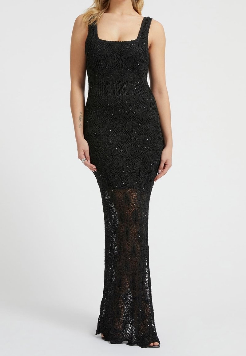 Nainen, joka on pukeutunut hihattomaan mustaan virkattuun mekkoon, jossa on neliönmuotoinen pääntie ja läpinäkyviä yksityiskohtia polvista lattiaan saakka.