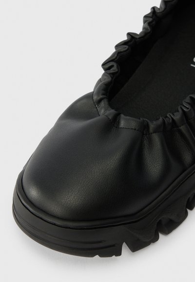 Zapato negro de cuero sin cordones con cuello elástico fruncido y suela texturizada y gruesa, mostrado en primer plano desde el lado frontal.