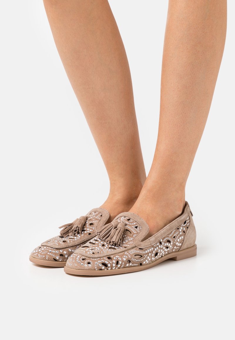 Mocassins en daim beige avec des découpes décoratives, des clous blancs et des glands portés pieds nus sur un fond blanc uni.