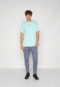 Hellblaues Baumwoll-T-Shirt mit kurzen Ärmeln und kleinem Logo auf der Brust; kombiniert mit grauen Jogginghosen mit Seitenstreifen und Logo.