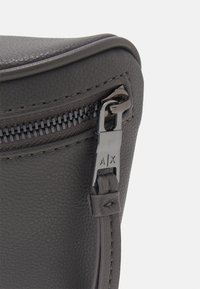 Róg szarej skórzanej torby z czarnym zamkiem błyskawicznym z wygrawerowaną metką z logo. Widoczna teksturowana powierzchnia oraz smukły, zaokrąglony kształt.