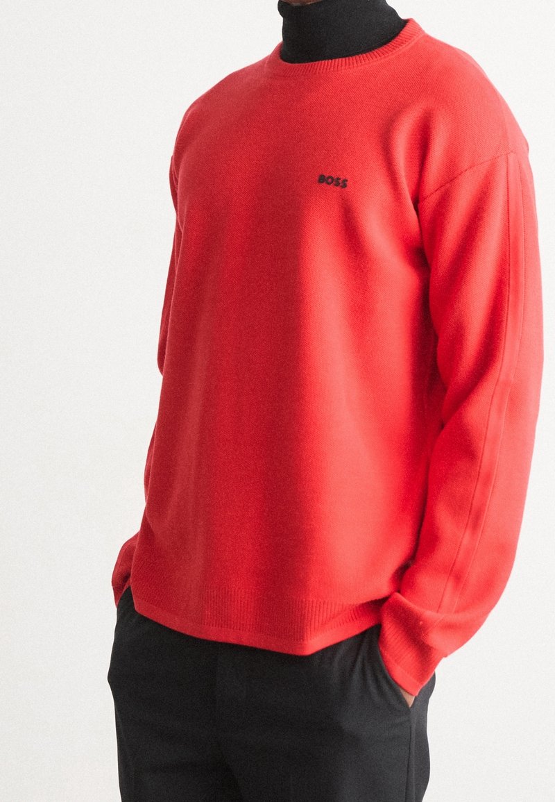Pull rouge à manches longues avec petit logo noir "BOSS" porté sur un col roulé noir, associé à un pantalon noir, mains dans les poches.