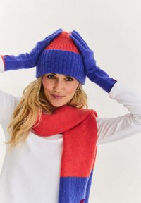 Pletená čepice, rukavice a šála v jednobarevných modrých a červených odstínech. Čepice má žebrovanou strukturu s trojúhelníkovým vzorem. Šála je rozdělena do barevných bloků.