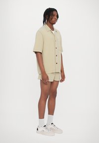 Chemise beige à manches courtes avec des boutons en bois, assortie à un short. Des chaussettes blanches et des baskets complètent la tenue, le tout se détachant sur un fond clair.