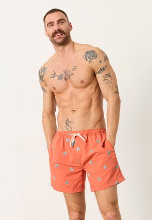 Skjorteløs mand med tatoveringer iført orange badeshorts med blå skildpadde-mønstre, hænderne i lommerne, smilende mod en ensfarvet lys baggrund.