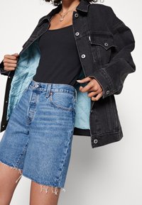 Giacca di denim nera con foderatura trapuntata blu chiaro, vestibilità oversized. Abbinata a pantaloni shorts di denim blu medio a vita alta, con dettagli di orlo sfrangiato.