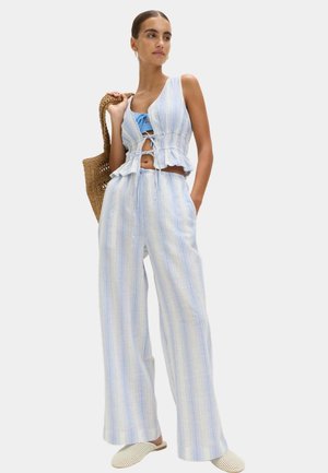 Frau trägt ein ärmelloses Oberteil mit hellblauen und weißen Streifen und weit geschnittene Hosen, hält eine gewebte Tasche und trägt weiße Slipper.