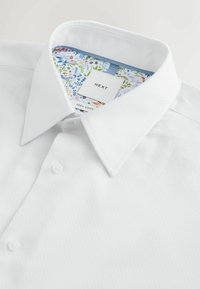 Biała bawełniana koszula z fakturowanej tkaniny, klasycznym kołnierzykiem, dwoma guzikami oraz kolorowym kwiatowym nadrukiem na wewnętrznej podszewce kołnierzyka.