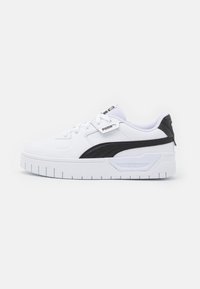 Sneaker blanche avec des accents noirs, présentant un dessus en cuir lisse, une pointe arrondie, une semelle épaisse avec un motif nervuré et des détails de marquage.