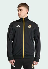 Czarna sportowa kurtka z żółtym zamkiem błyskawicznym, z białym logo Adidas i emblematem Realu Madryt. Wykonana z gładkiego, lekkiego materiału, z długimi rękawami.