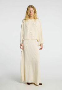 Conjunto de dos piezas color crema que incluye una blusa de manga larga y una falda maxi, ambos con costuras diagonales y fabricados en una tela suave y fluida.