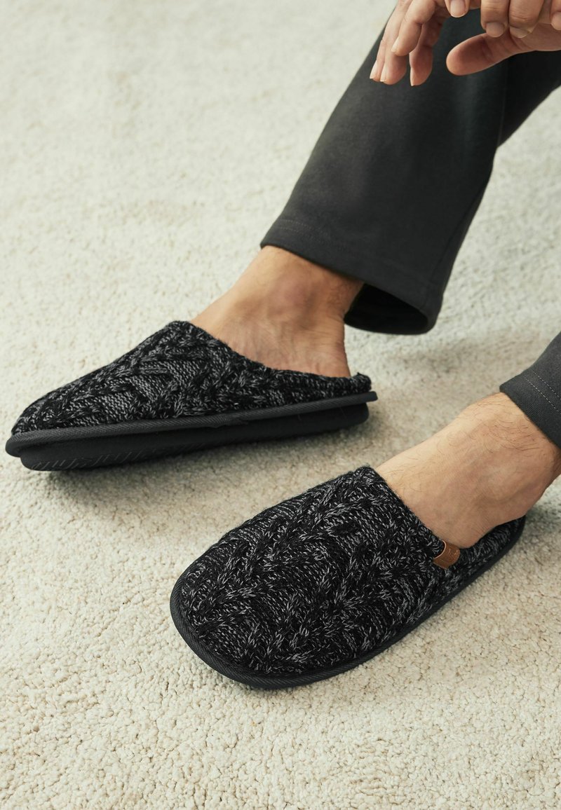 Black knitted slippers with a textured pattern, featuring a black rubber sole and a small brown accent label, worn on bare feet.