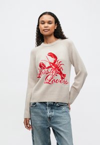 Mujer con cabello rizado vistiendo un suéter beige con un gran langosta roja y el texto "Amantes de las Langostas", combinado con jeans azules descoloridos, contra un fondo blanco.