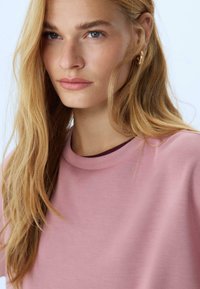 Donna con lunghi capelli biondi indossa una maglietta rosa chiaro a girocollo e orecchini a cerchio dorati, guarda leggermente di lato contro uno sfondo chiaro.