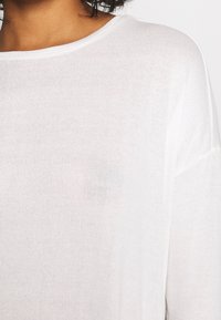 Närbild av en person som bär en mjuk vit stickad tröja med rund halsringning och något sänkta axlar.