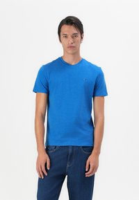 Blaues Baumwoll-T-Shirt mit Rundhalsausschnitt und kurzen Ärmeln. Hat ein kleines Logo auf der Brust. Kombiniert mit dunkelblauen Jeans.
