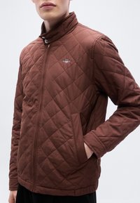 Καφέ καπιτονέ τζάκετ με ψηλό γιακά, διαθέτει εμπρόσθιο φερμουάρ και πλαϊνές τσέπες. Μικρή λεπτομέρεια λογοτύπου στο πάνω αριστερό μέρος.