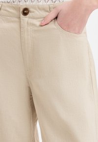 Beige broek met een gladde, lichte textuur, voorzien van een zijzak en een knoopsluiting. Eenvoudig ontwerp, op maat gemaakte pasvorm zichtbaar bij de heup.