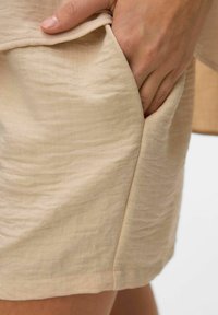 Jasno-beżowe szorty z tkaniny o teksturowanym wykończeniu i boczną kieszenią. Szwy są starannie wykończone, a materiał układa się miękko.