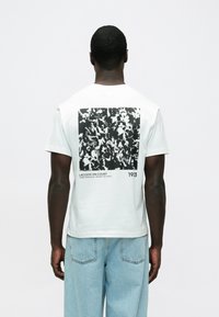 Biała bawełniana koszulka z czarno-białym nadrukiem na plecach, z napisem "LACOSTE ON COURT 1933" i kwadratowym wzorem.