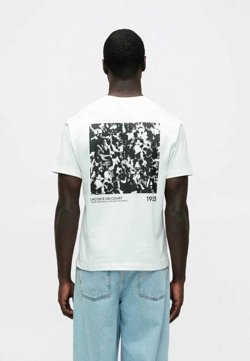 Biała bawełniana koszulka z czarno-białym nadrukiem na plecach, z napisem "LACOSTE ON COURT 1933" i kwadratowym wzorem.
