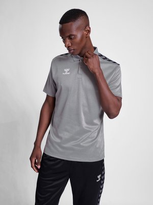 Mann richtet den Kragen eines grauen Hummel Kurzarm-Poloshirts, trägt schwarze Hosen mit Hummel-Logo vor schlichtem hellem Hintergrund.