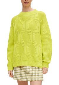 Limegrüner Strickpullover mit geometrischem Muster, geripptem Rundhalsausschnitt und überschnittenen Schultern. Getragen über einem karierten Rock.