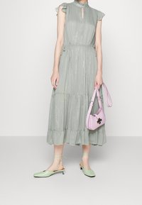 Kvinna iklädd en skimrande ljusgrön midiklänning, håller en pastellrosa handväska och bär matchande ljusgröna lågklackade skor med ankeldrag.