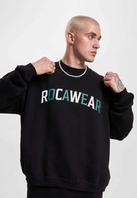 Mustat collegepuserot, joissa on "ROCAWEAR"-logo valkoisena ja turkoosina, ribbireunat kaulassa ja hihoissa, rento istuvuus, pehmeä materiaali. Malli säätää vaatetta.