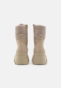 Botas de tobillo impermeables en color beige con un eje acolchado, cremallera lateral y suela gruesa texturizada. La parte superior combina materiales lisos y acolchados.