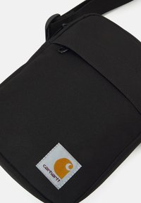 Črn krčkovni nahrbtnik z zadrgo, ki ima sivo nalepko z oranžnim logotipom in pravokotno obliko. Teksturirana površina.