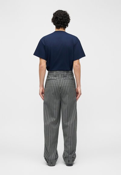 Άτομο με σγουρά μαλλιά που φοράει μπλε μπλουζάκι και φαρδιά γκρι παντελόνια με κάθετες ρίγες, στέκεται γυρισμένο πλάτη σε απλό φόντο.