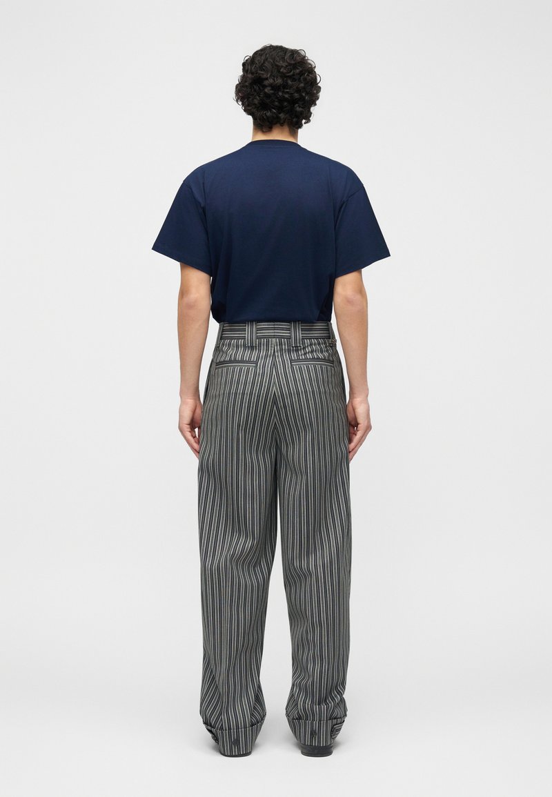 Persona con capelli ricci che indossa una maglietta blu navy e pantaloni larghi grigi con strisce verticali, vista di schiena su uno sfondo neutro.