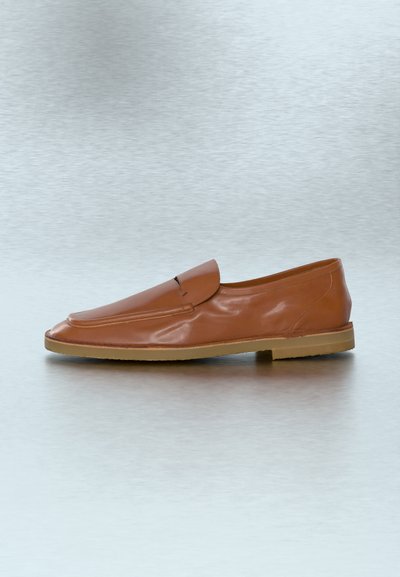Chaussure marron en cuir à enfiler, avec une semelle en caoutchouc plate, un design épuré et des détails de couture minimalistes sur un fond neutre.