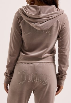 Donna che indossa una felpa con cappuccio e pantaloni color taupe in velluto con loghi "Juicy" tempestati di strass sulla schiena della maglia e dei pantaloni, vista di spalle.