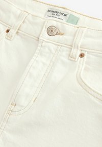 Mittellange Boyfriend-Shorts aus hellcremfarbener Jeans. Mit leichtem Stich, einem metallischen Knopfverschluss und einem Etikett, das Größe und Marke anzeigt.