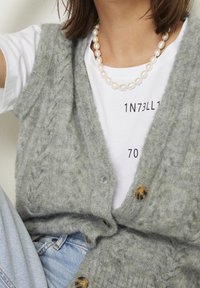 Grå stickad väst med flätmönster, prydd med stora bruna knappar, lagd över en vit t-shirt med tryckt text, tillsammans med blå jeans.