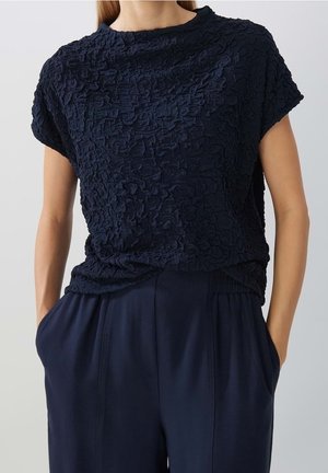 Marineblå tekstureret kortærmet top med blomsterprint parret med løstsiddende marineblå bukser, model med hænderne i lommerne mod ensfarvet baggrund.