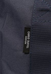 Borsa a duffel blu navy realizzata in tessuto testurizzato, con etichetta nera e testo bianco "CARGO DUFFEL MEDIUM." Dettagli cuciti visibili.