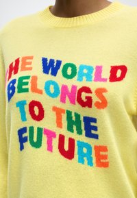 Κίτρινο πουλόβερ με πολύχρωμο κείμενο μπροστά που λέει «Ο ΚΟΣΜΟΣ ΑΝΗΚΕΙ ΣΤΟ ΜΕΛΛΟΝ».