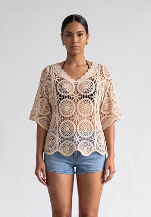 Donna che indossa un top beige all'uncinetto con motivi circolari sopra un top bikini nero e pantaloncini di jeans blu, in piedi davanti a uno sfondo semplice.