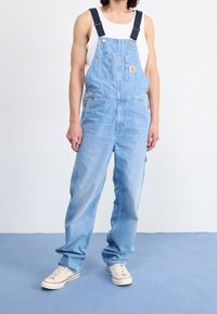 Helle blaue Jeansoverall mit verstellbaren dunklen Trägern, Fronttaschen und Messingbeschlägen. Kombiniert mit einem weißen Tanktop und Turnschuhen.