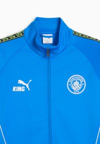 Sininen urheilu takki, joka on valmistettu sileästä kankaasta. Takissa on korkea kaulus, täysi vetoketju, valkoiset sivuakkentsit ja painetut logot: Puma, KING, Manchester City.