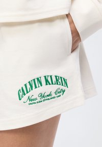 Χέρι στην τσέπη λευκών σορτς με πράσινο λογότυπο «Calvin Klein New York City» κεντημένο κοντά στο τελείωμα.