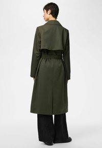 Lühikeste tumedate juustega inimene kannab pikkade vööga oliivirohelise värvi trentsi, musti laiade sääreosadega pükse ja musti kingi, seisab seljaga vaataja poole.