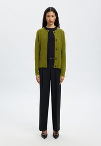 Cárdigan verde de punto con escote redondo, cierre de botones, combinado con una camiseta negra y pantalones negros a medida, junto con zapatos negros.